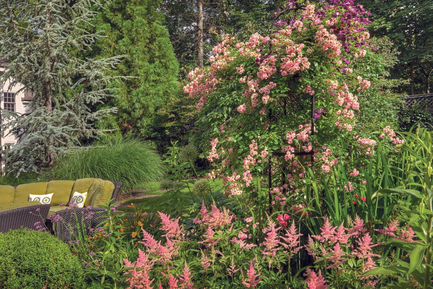 A lush garden with diverse plants, colorful flowers, a wicker sofa with cushions, and a backdrop of various green trees and shrubs.