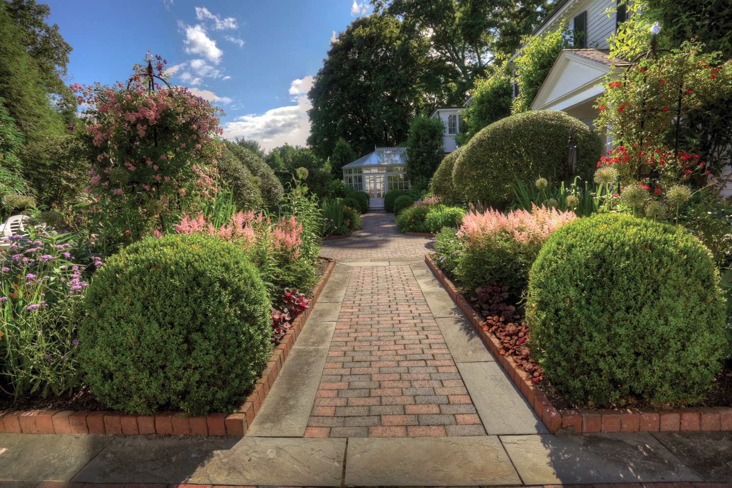 A brick path leads to a white greenhouse, flanked by manicured bushes and vibrant flower beds, under a partly cloudy sky in a lush garden.