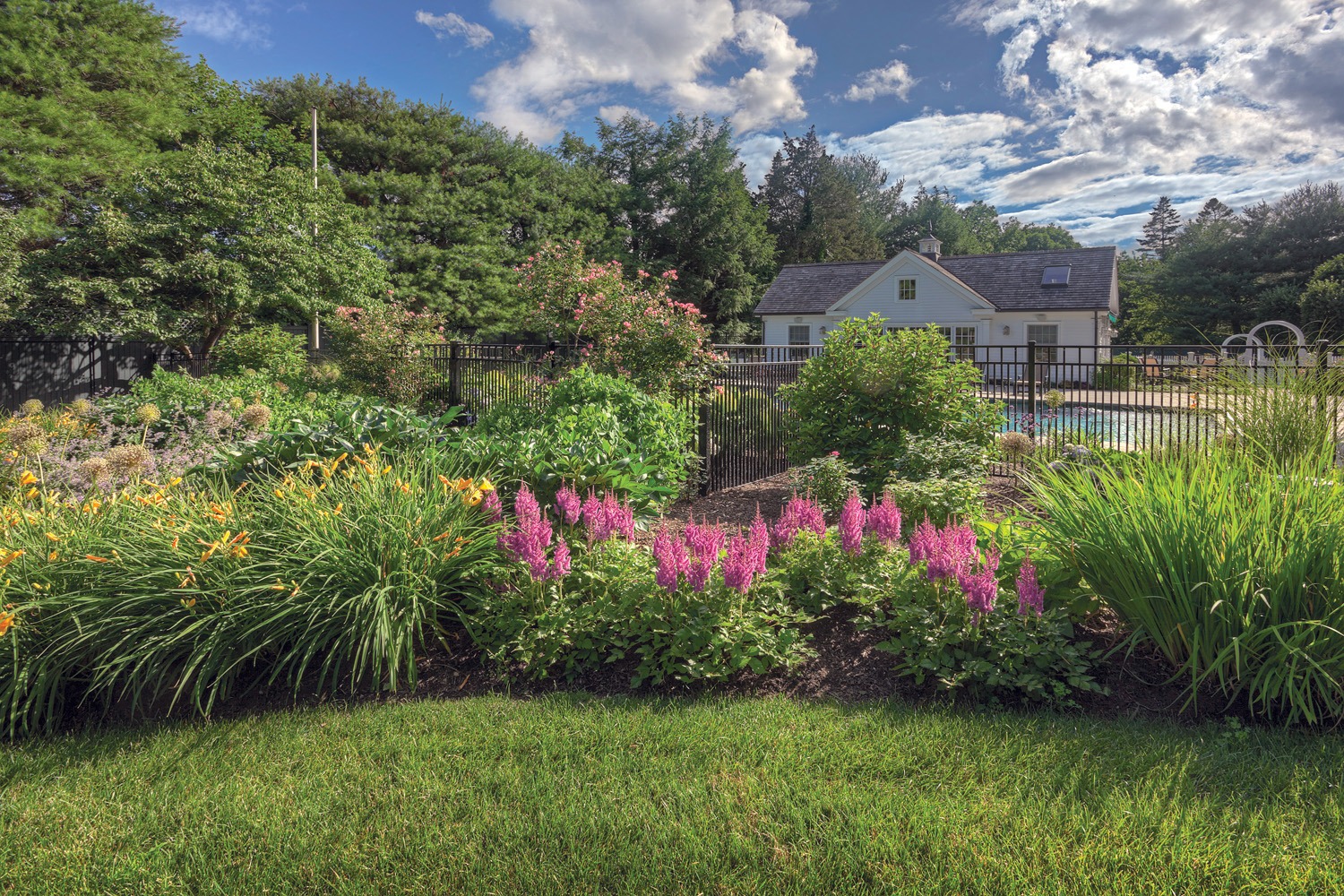 A well-manicured garden with vibrant flowers in front, a pool behind a black fence, and a small house with white walls and a shingled roof.