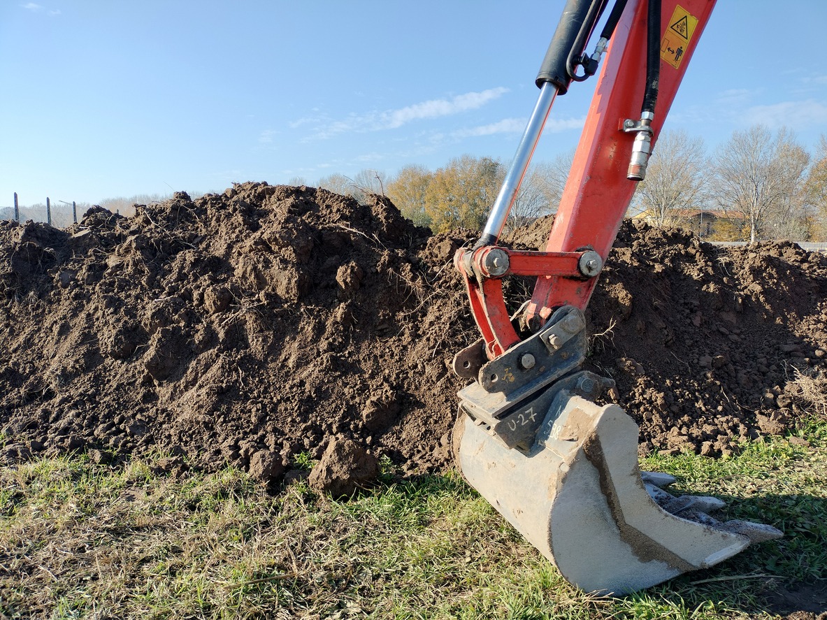 A close-up of an excavator bucket at a construction site with a large pile of freshly dug earth in the background under a clear blue sky.