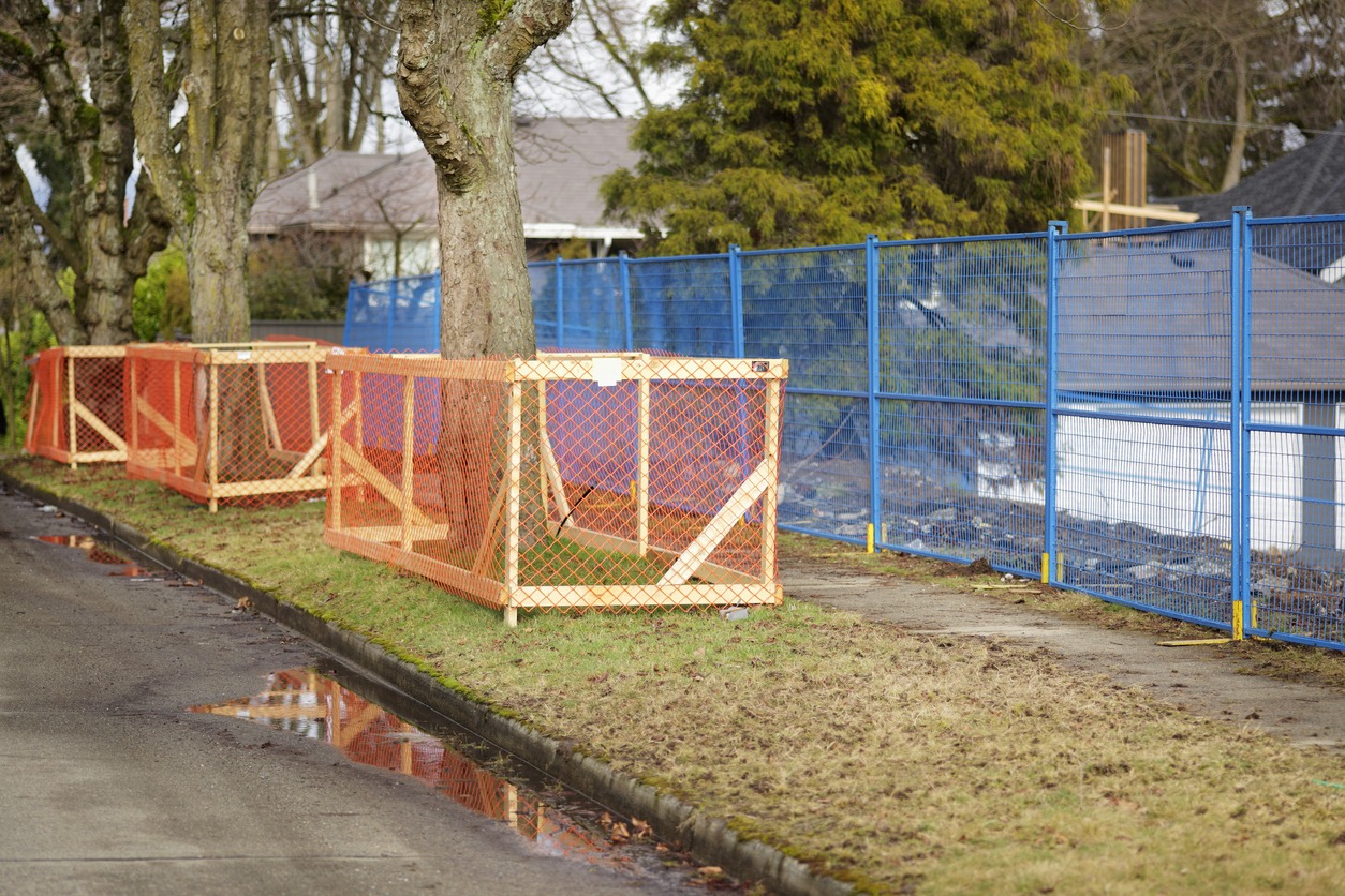 A fenced-off construction site with orange and blue barriers along a wet sidewalk, beside leafless trees, with a residential backdrop.