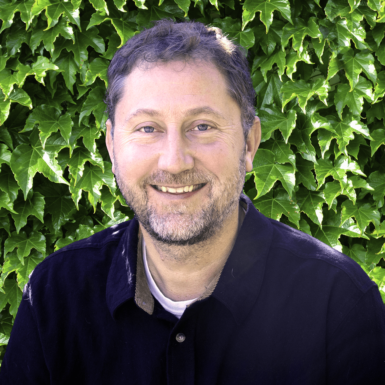 This image shows a smiling person with short graying hair, wearing a dark jacket, in front of a lush green ivy background.